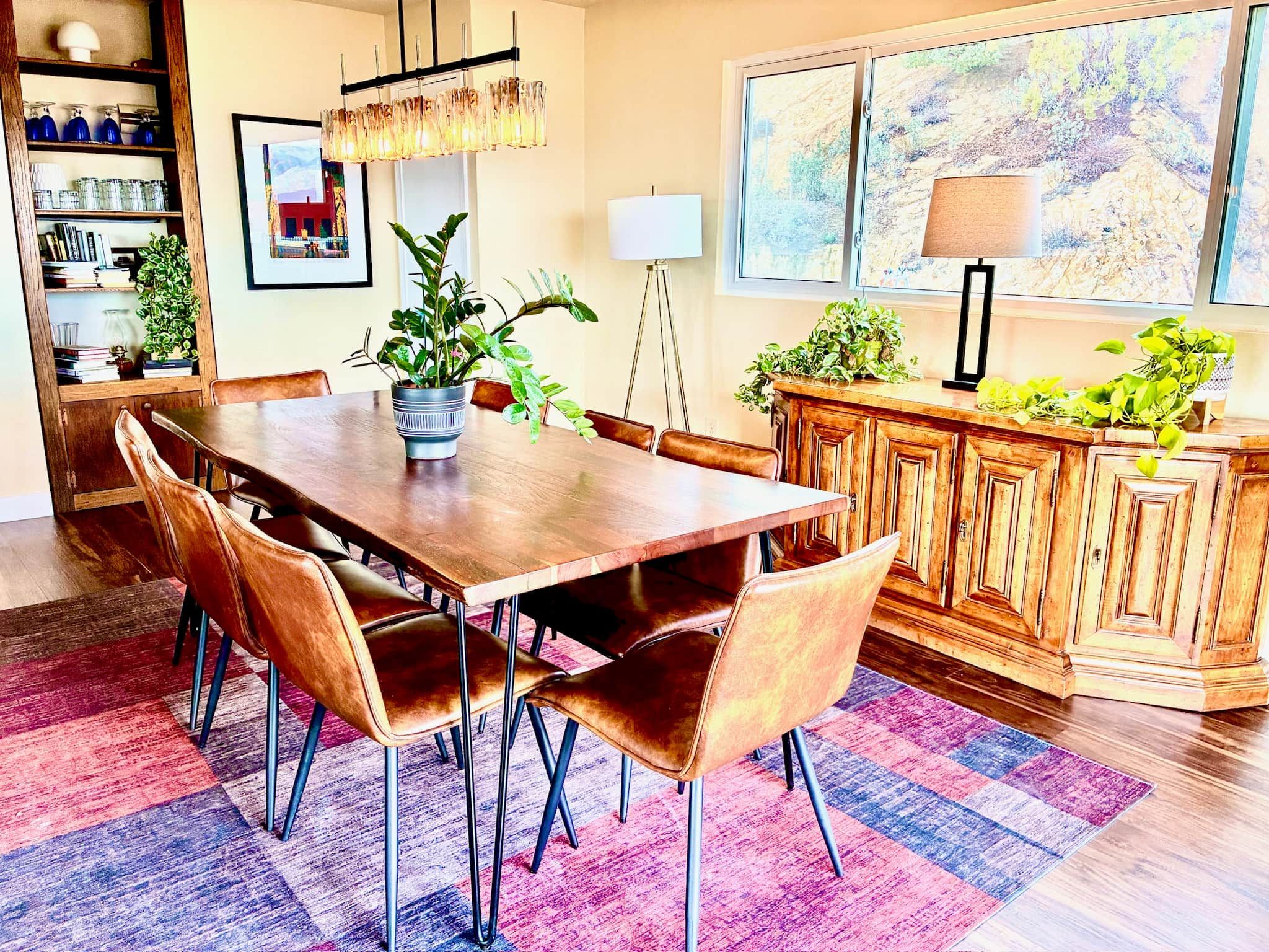A dining area with a wooden table, leather chairs, a potted plant, and a colorful rug.
