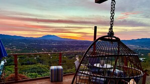 Ein Hängekorbstuhl mit Kissen auf einer Holzterrasse mit Blick auf eine Landschaft, Berge und einen Sonnenuntergang.