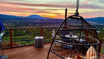 A hanging chair with cushions on a wooden deck overlooking a landscape with mountains and a sunset.