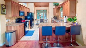 A modern kitchen with wooden cabinets, a stainless steel refrigerator, a microwave, and a red stove. There are two bar stools and a blue rug.