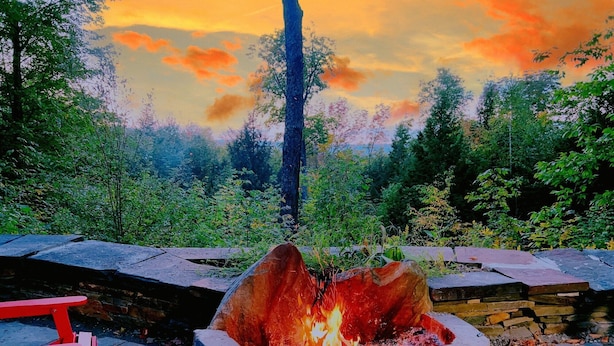 A fire pit surrounded by stone seating, set in a forested area with a view of a sunset.