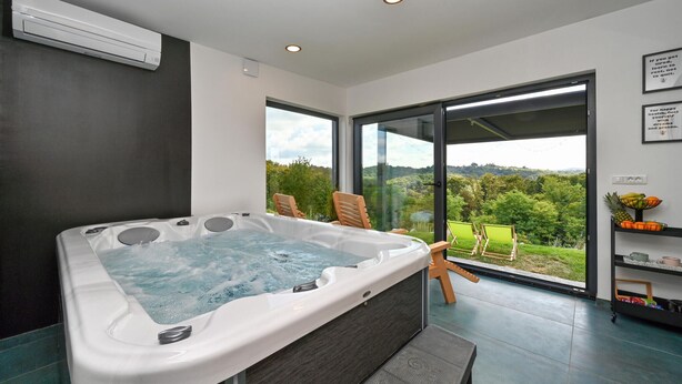 A modern bathroom with a large bathtub, a shower area, and a view of a lush landscape through large windows. - Luxurious home in Banfi with sauna (Banfi)