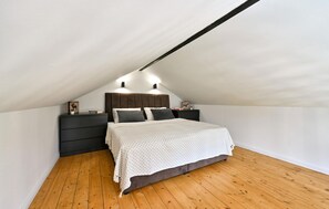 Een slaapkamer met een schuine plafond, een bed met een wit beddegoed met patroon, twee nachtkastjes met lampen, en een houten vloer.