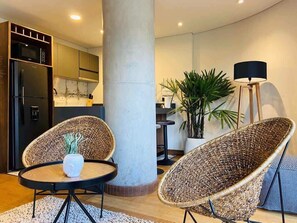Una sala moderna con sillas de mimbre, una mesa de centro blanca, un pilar grande y una lámpara de pie.