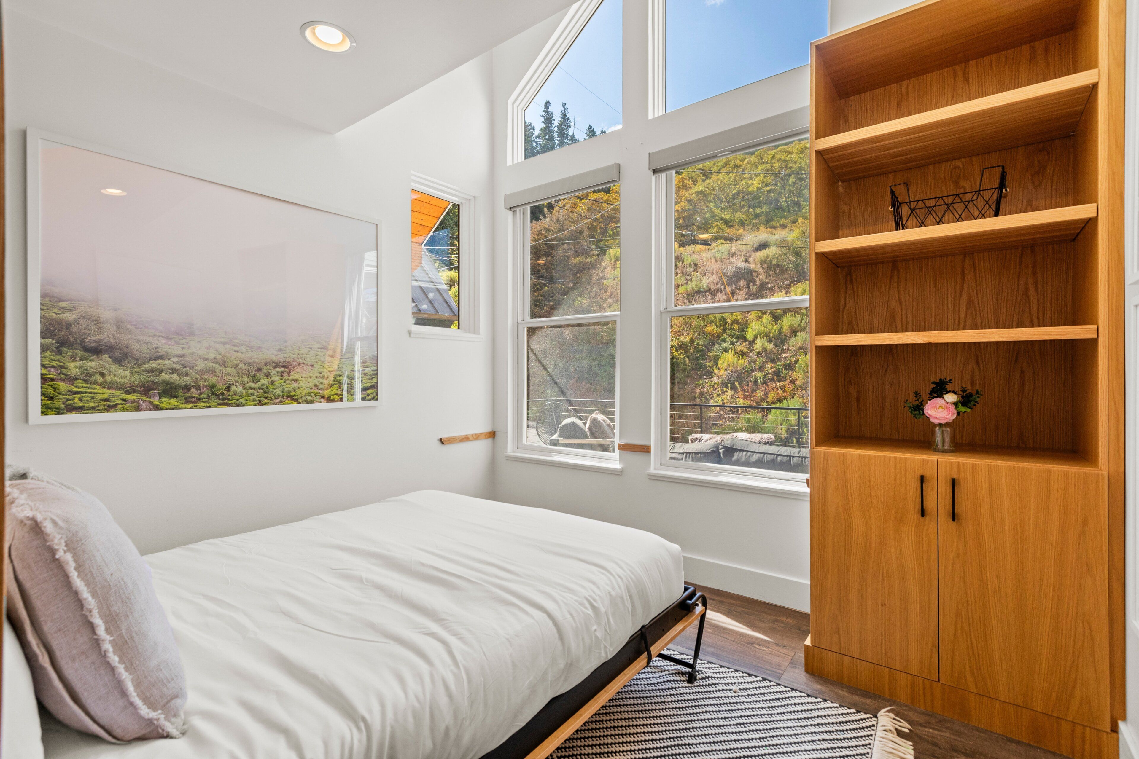 A bedroom with a bed, a wooden bookshelf, and large windows.