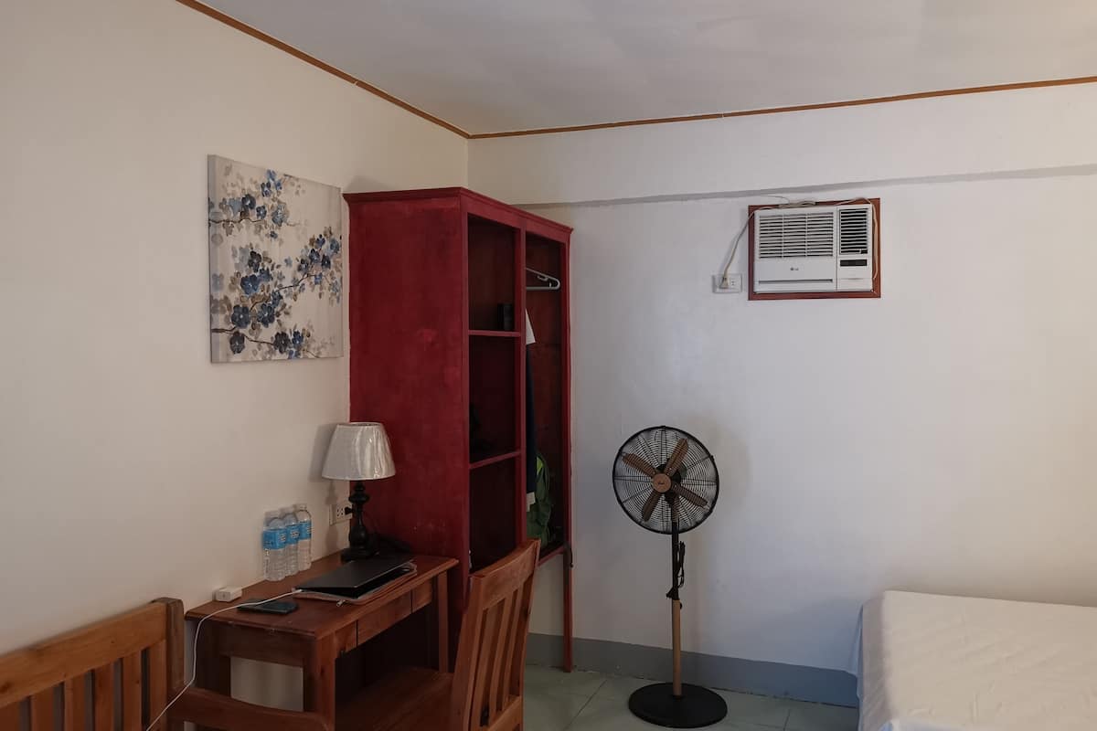A room with a wooden desk, a red cabinet, a standing fan, and a bed.