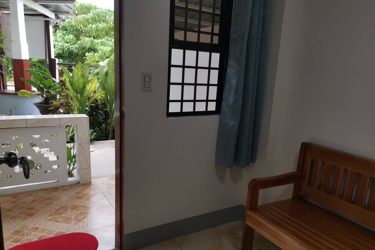 A room with a wooden bench, a red doormat, a white door with a window, and a view of greenery outside.