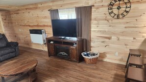 A cozy living room with a wooden floor, a television on a wooden stand, a fireplace, a clock on the wall, and a wicker basket.
