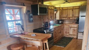 A wooden cabin interior with a kitchen area featuring a stove, sink, and cabinets. There are wooden stools around a table and a window with a view of a red structure outside. - Charming 2-bedroom cabin in Carey with AC, WiFi (Carey)