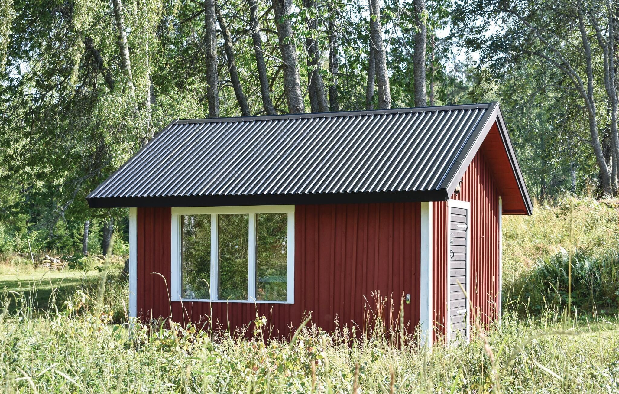 Une petite cabane en bois rouge, avec un toit métallique incliné, entourée de grands arbres et de hautes herbes.
