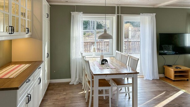 Un coin repas avec une table et des chaises en bois, un luminaire suspendu et une vue sur l’extérieur grâce à de grandes fenêtres.