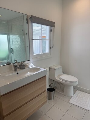 Un baño con un lavamanos blanco, un inodoro, un espejo y una ventana con persiana blanca.