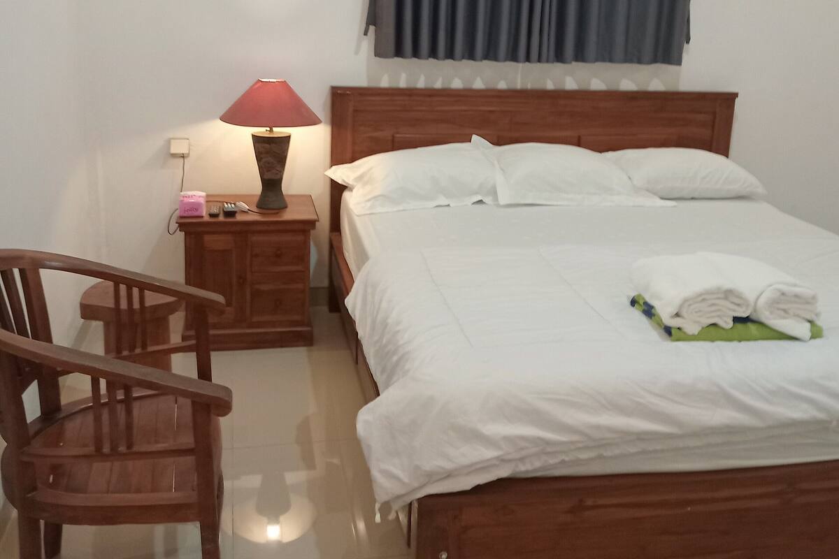 Una cama bien hecha con ropa blanca, un cabecero de madera y una mesita de noche con una lámpara y un objeto rosa.