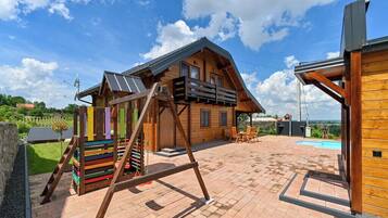 Une cabane en bois avec un toit en pente, une terrasse avec une piscine et une aire de jeux colorée.