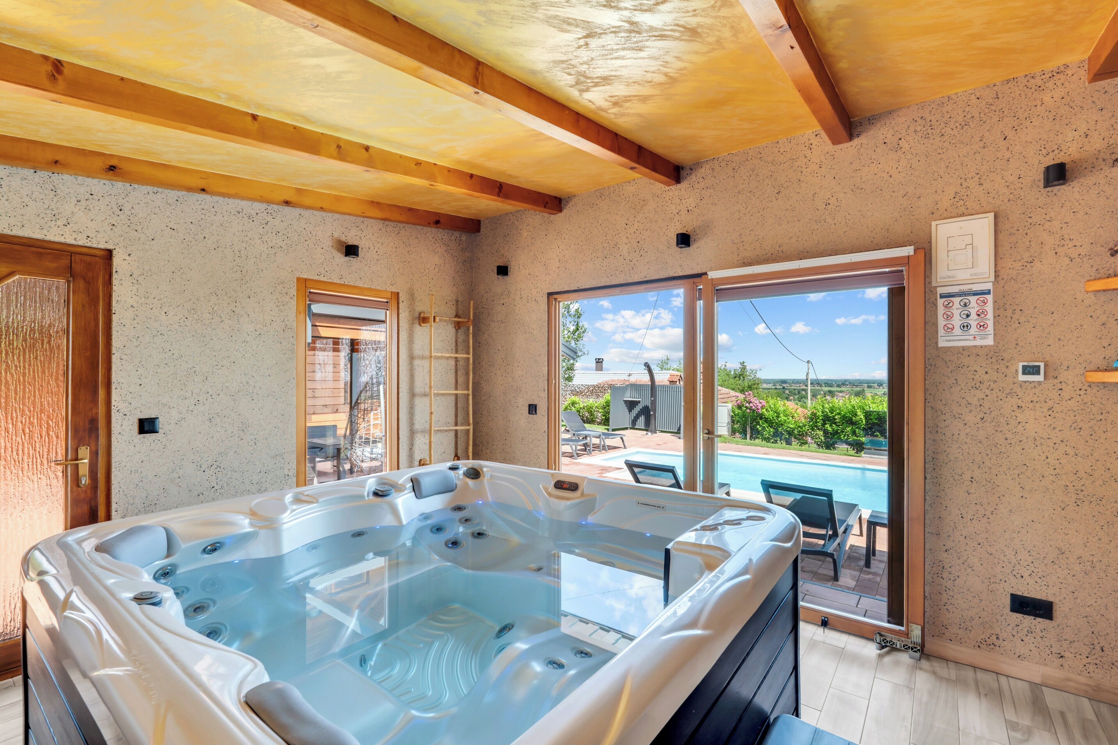 Ein Wellnessraum mit Whirlpool, Holzbalkendecke und Blick auf Schwimmbad und Garten.