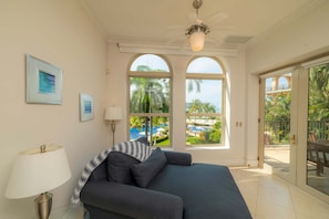 A room with a sofa, a lamp, and large windows with a view of a pool and palm trees.