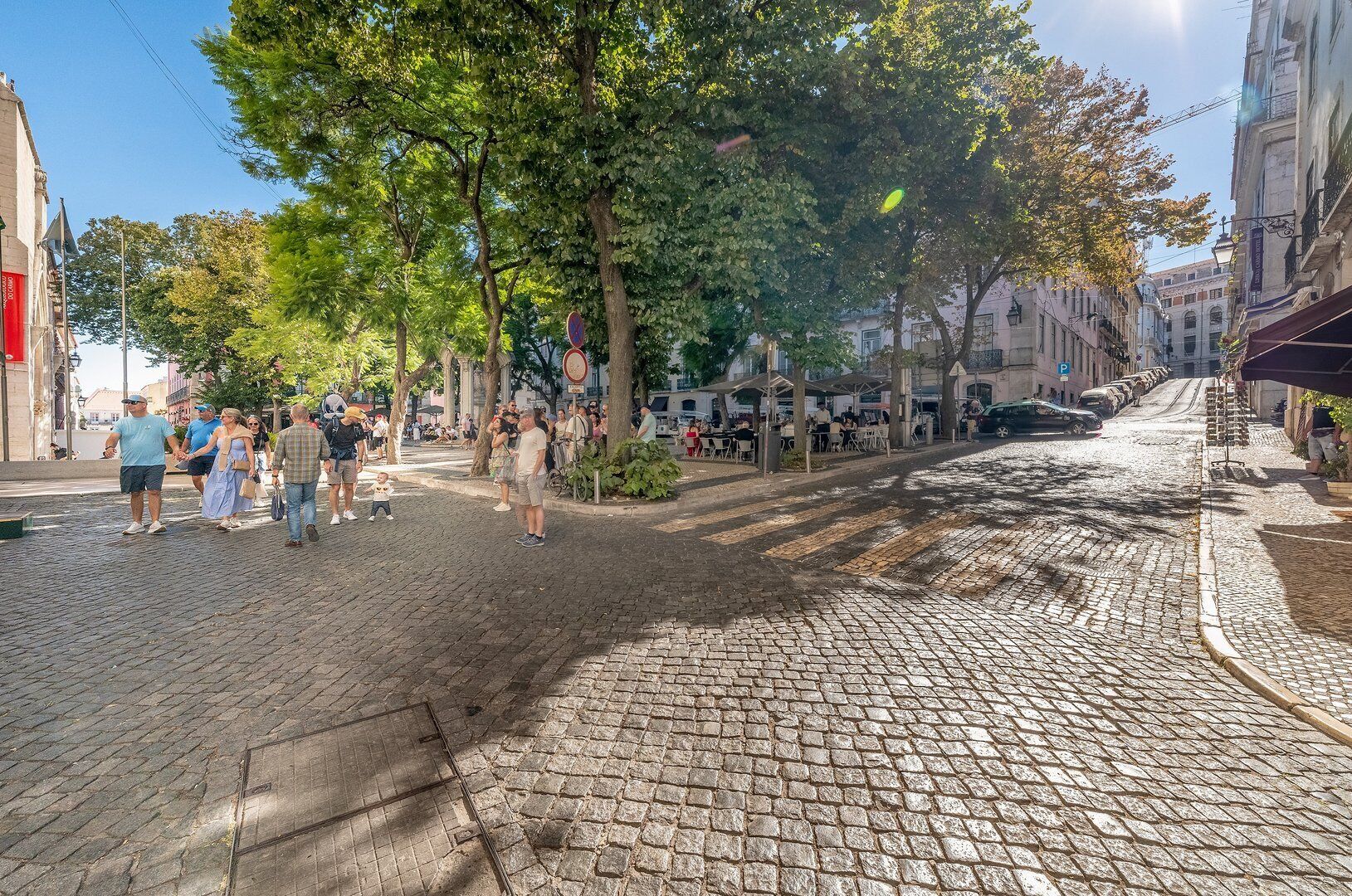 Une rue pavée bordée d’arbres et de bâtiments, où l’on voit des gens se promener et s’asseoir aux terrasses des cafés.