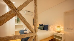 A bedroom with a wooden ceiling structure, a bed with pillows, a bedside table with a lamp, and a small shelf with towels.