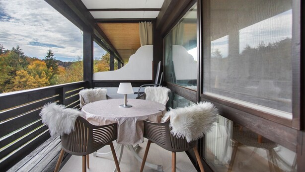 Een balkon met een ronde tafel, stoelen met zachte kussens en uitzicht op herfstbomen.