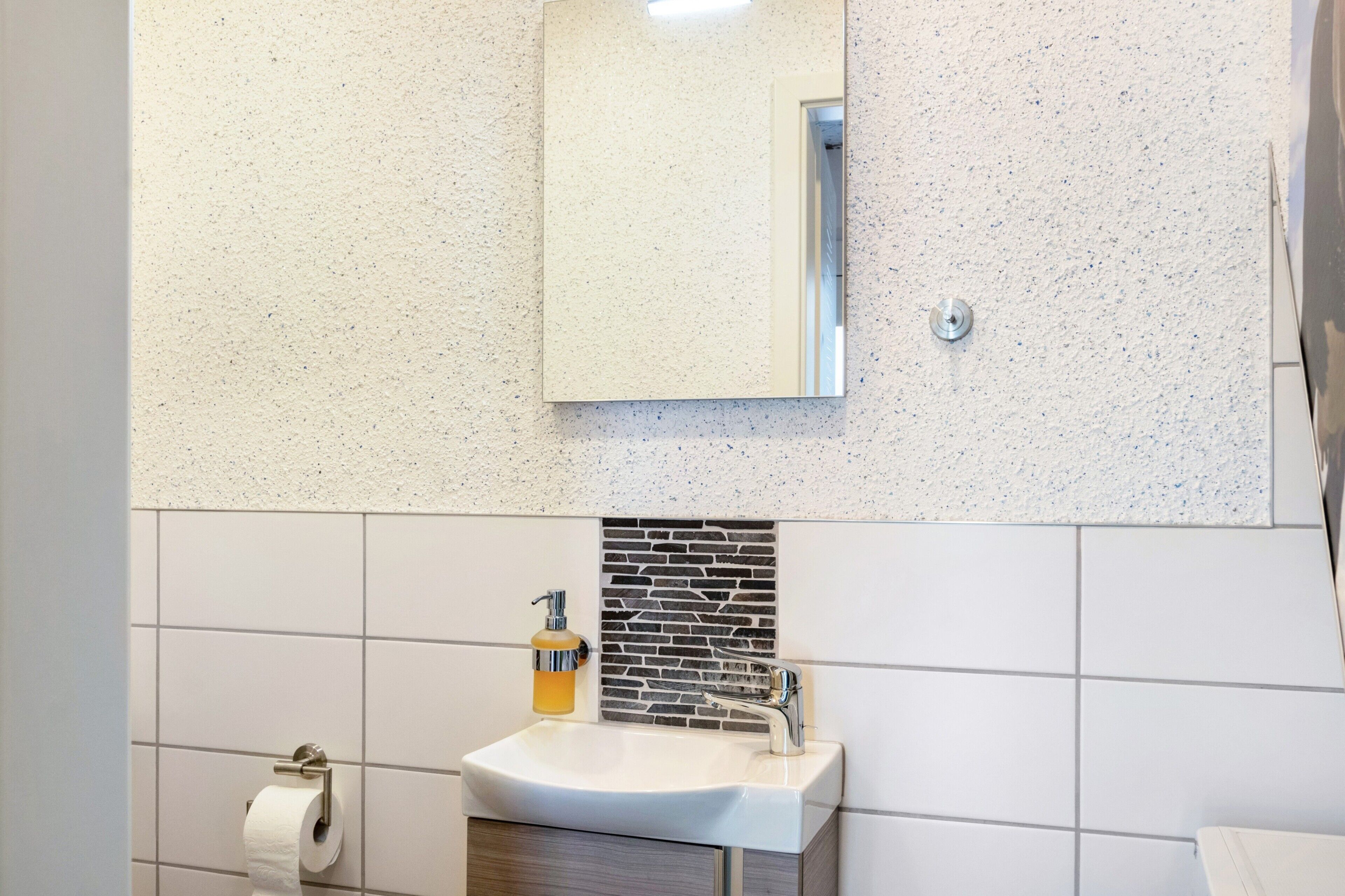 A bathroom with a white sink, a mirror, a soap dispenser, and a toilet paper holder.