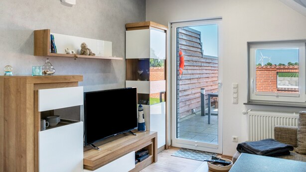 A modern living room with a flat-screen TV, wooden shelving, a sliding glass door to an outdoor area, and a window with a view of wind turbines. - Apartment 'Fewo Kaso' with Private Garden, Wi-Fi and Air Conditioning (Ihlow)