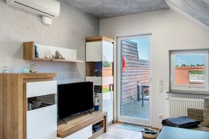 Ein modernes Wohnzimmer mit Flachbildfernseher, Holzregalen, einer Glastür zum Außenbereich und einem Fenster mit Blick auf Windkraftanlagen.