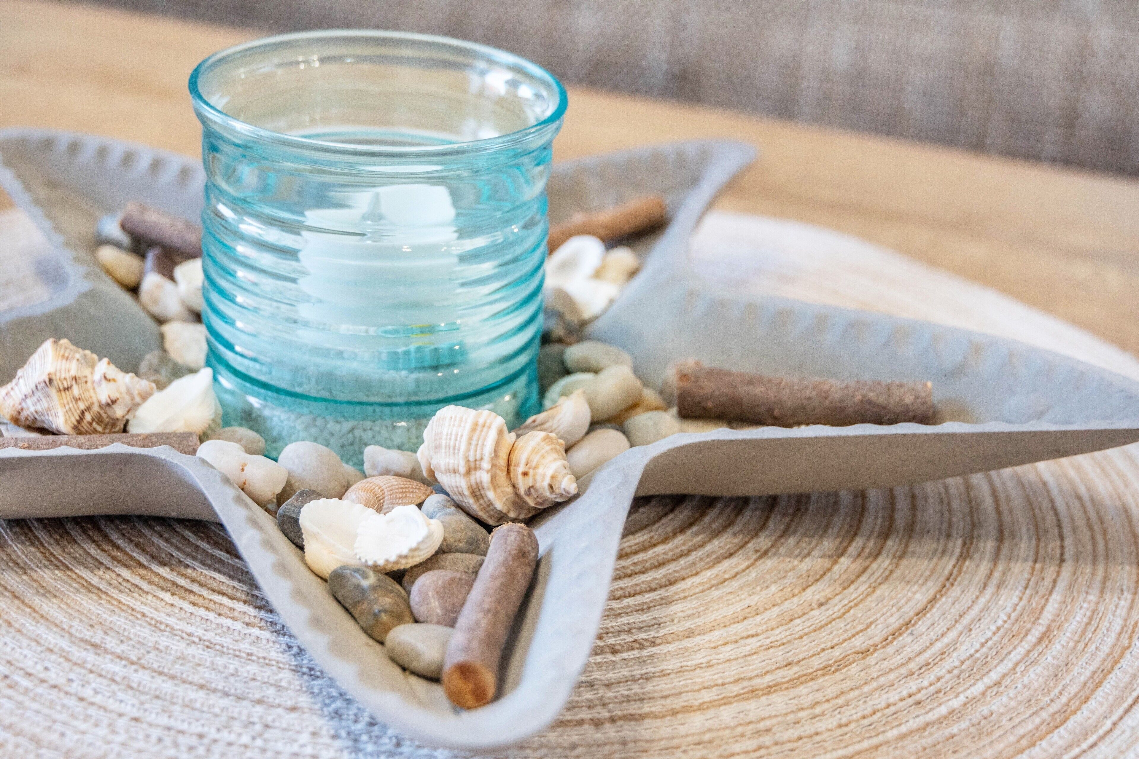 A glass container with a blue tint surrounded by various seashells and smooth stones on a textured surface.