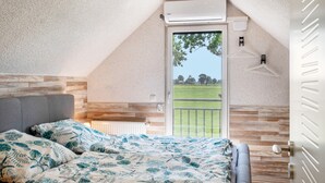 A bedroom with a sloped ceiling, a bed with patterned bedding, a radiator, a window with a view of greenery, and a door with a handle. - Apartment 'Fewo Kaso' with Private Garden, Wi-Fi and Air Conditioning (Ihlow)