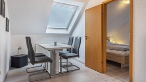 Een kleine kamer met dakraam, een eettafel met stoelen, een nachtkastje met lamp en een bed.
