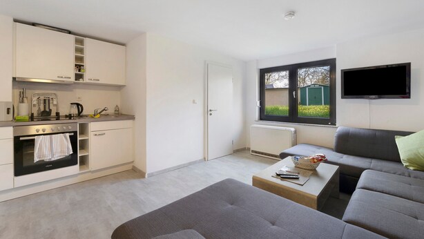 A modern kitchen with built-in appliances, a dining area with a grey sofa and a table, and a TV mounted on the wall. - New Central Apartment with Wi-Fi in Stuttgart (Stuttgart)