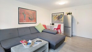 A modern living room with a grey sofa, a small coffee table with snacks and a remote, a dining area with a white table and red chairs, and a refrigerator with wine bottles. - New Central Apartment with Wi-Fi in Stuttgart (Stuttgart)