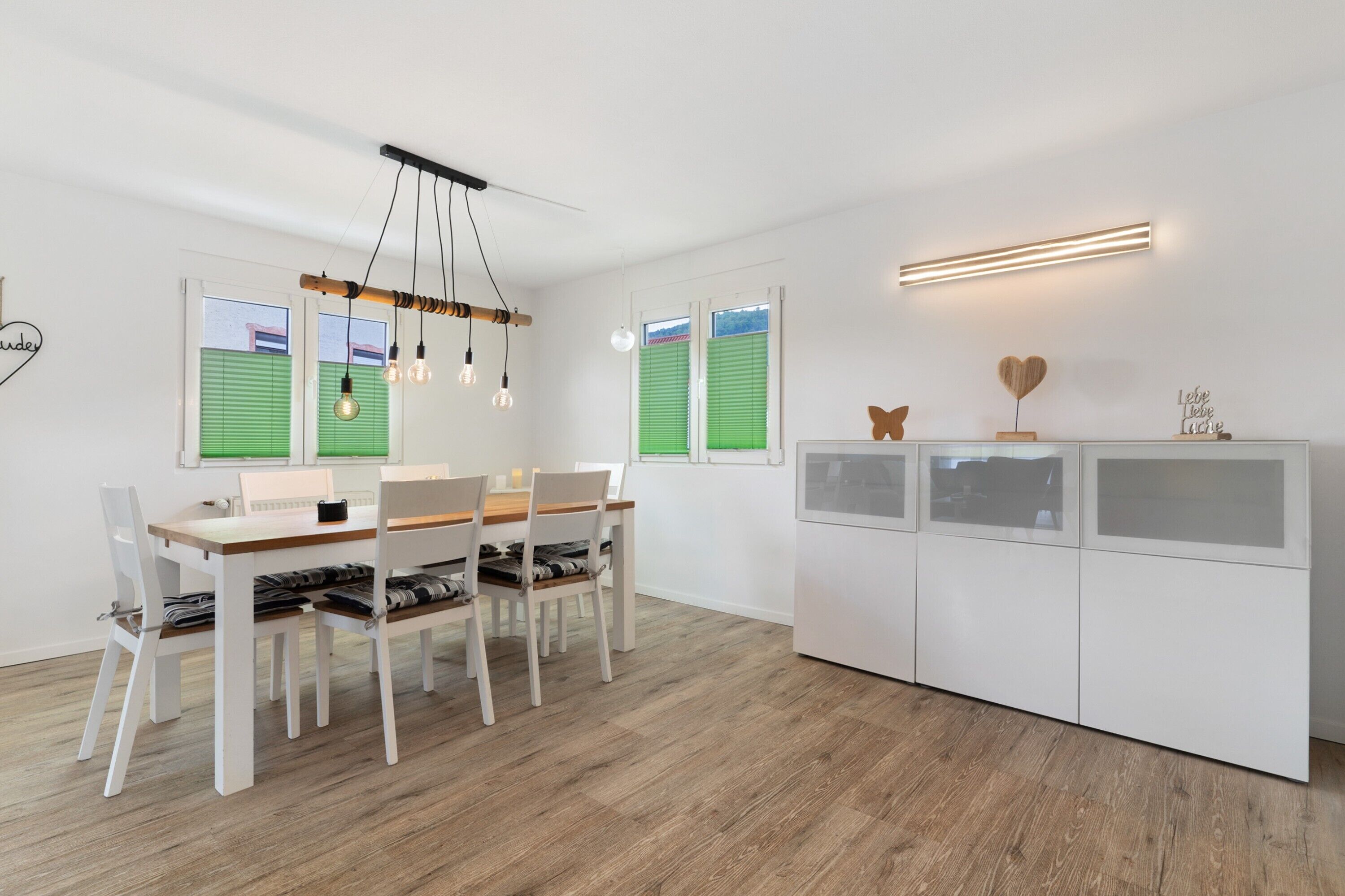 Un comedor moderno con una mesa y sillas de madera, una lámpara colgante y un armario blanco con adornos.