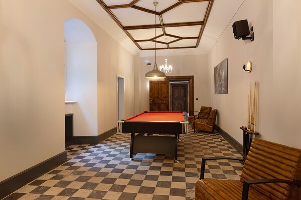 A hallway with a pool table, checkered floor, wooden chairs, a chandelier, and a door with a wooden frame.