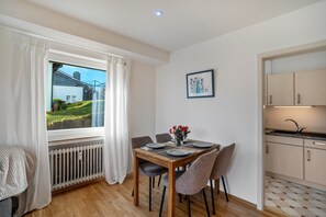Un coin repas avec une table dressée pour quatre personnes, un radiateur, une fenêtre avec des rideaux et un espace cuisine avec des placards.