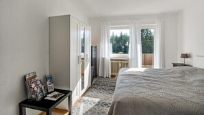 Ein Hotelzimmer mit Bett, Kleiderschrank, Tisch mit Zeitschriften, Fenster mit Vorhängen und Blick auf Bäume.