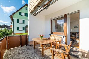 Une terrasse avec une table et des chaises en bois, un petit vase avec des fleurs et une porte-fenêtre coulissante donnant sur un espace intérieur.