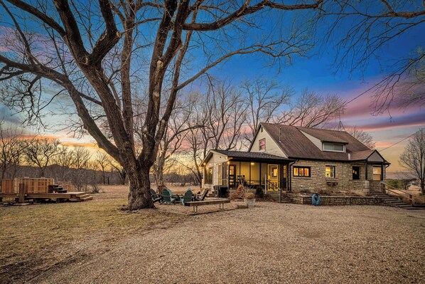 A stone house with a porch, outdoor seating, and a large tree in front.