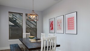 Un comedor con una mesa de madera, sillas blancas, un lámpara colgante y tres cuadros en la pared.