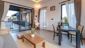 A modern living room with a wooden coffee table, a sofa, and a dining table with chairs. There is a balcony with a glass door and a view of the outside. - Villa Blessing Bali (Ungasan)