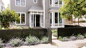 A two-story house with a front porch, a picket fence, and a garden with purple flowers.
