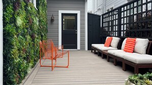 A modern outdoor patio with a wooden deck, a black door, a seating area with white cushions and orange pillows, and a vertical garden on the left.