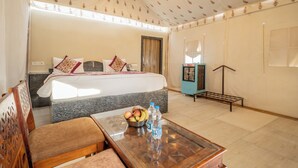Una habitación de hotel con una cama, una pequeña mesa con fruteros y botellas de agua, una zona de estar con un sofá y un ventanal grande.