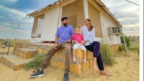 Una familia de cuatro personas sentada frente a una casa pequeña de una sola planta, con cimientos de piedra y un techo a dos aguas sencillo.