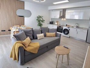 Una sala moderna con un sofá gris, una mesita redonda pequeña y un comedor con mesa y sillas de madera. Se ve una lavadora en la cocina.