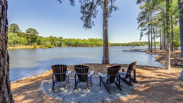 Een zitgedeelte aan het meer met drie Adirondack-stoelen en een houten bank, omringd door bomen en een helder uitzicht op het water.