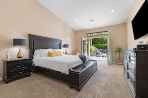 Quarto com cama grande, cabeceira de madeira escura, duas mesas de cabeceira com luminárias, um banco ao pé da cama, cômoda e televisão na parede.