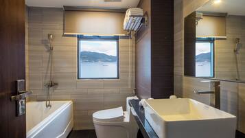 A modern bathroom with a bathtub, toilet, and sink, featuring a large window with a view of the sea.