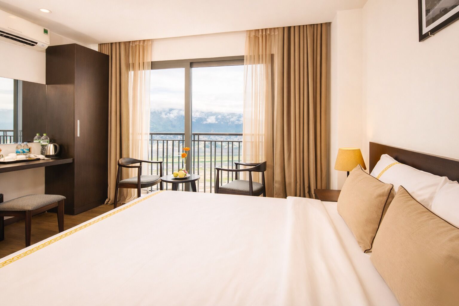 A hotel room with a large bed, a desk with a chair, a small table with a chair, and a balcony with a view.