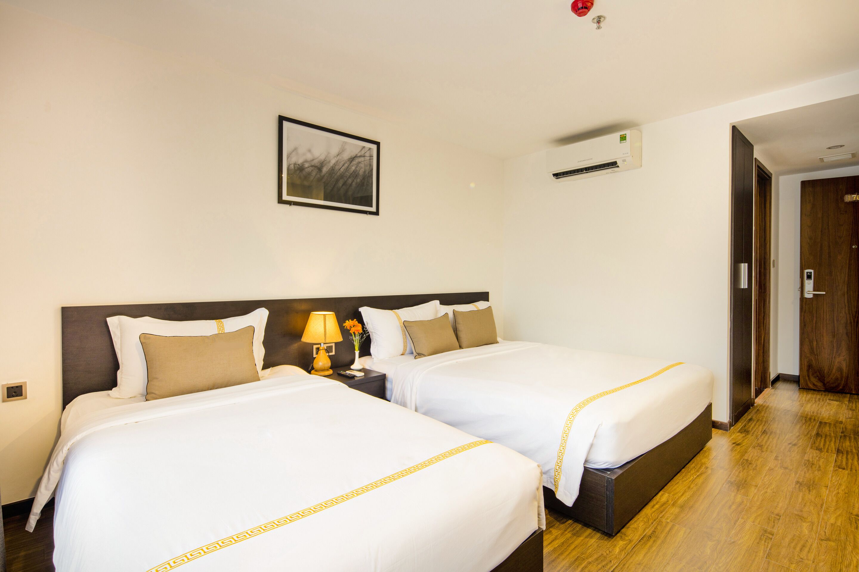 A hotel room with two beds, a nightstand with a lamp, a framed picture on the wall, and an air conditioning unit.