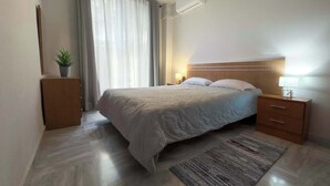 A modern bedroom with a large bed, a bedside table with a lamp, a small plant, and an air conditioning unit on the wall.